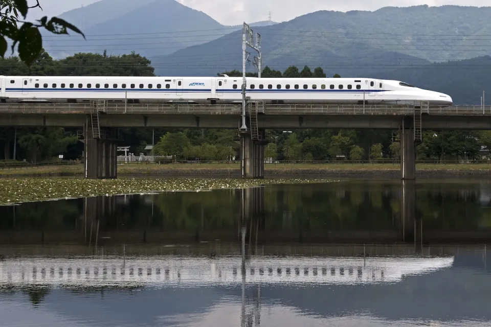 White Shinkansen high-speed rail train passing on bridge over pond in Japan.